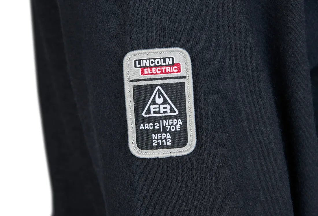 A close-up of the Lincoln K5267-XL Arc Rated FR Welding T-Shirt XLarge black sleeve shows a rectangular patch with the Lincoln Electric logo and safety certification labels for arc rating, NFPA 70E, and NFPA 2112.