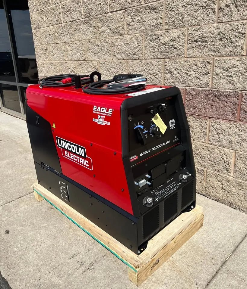 A Lincoln Eagle 10,000 Plus Engine Drive Factory Demo Welder (U2343-4) in red and black sits on a wooden pallet outside, near a tan brick wall and glass window.