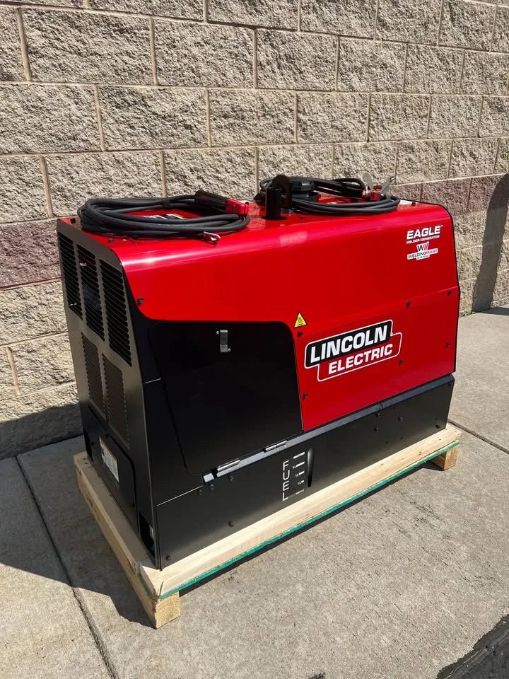 A Lincoln Eagle 10,000 Plus Engine Drive Factory Demo Welder (U2343-4) in red and black rests on a wooden pallet outside by a tan brick wall on a concrete sidewalk, cables coiled on top.