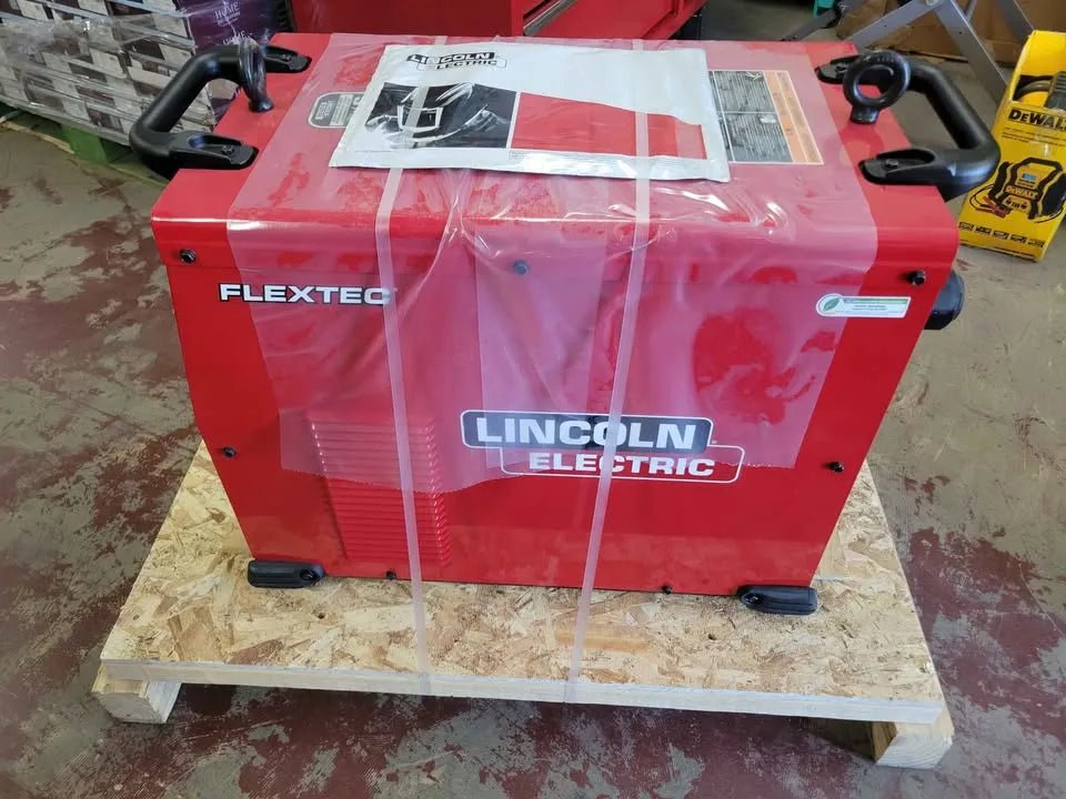 A Lincoln Flextec 650X Multi-Process Factory Demo Welder with CrossLinc Technology (U3425-1) in red sits on a wooden pallet, secured with plastic wrap. A manual is on top, and the worn concrete floor is visible beneath.