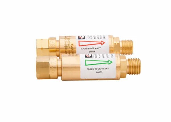 Two brass-colored cylindrical adapters, labeled Made in Germany, serve as regulator-type flashguards for oxygen and fuel gas. Each has threaded ends and a colored arrow—red or green—to indicate flow direction for these flashback arrestors.