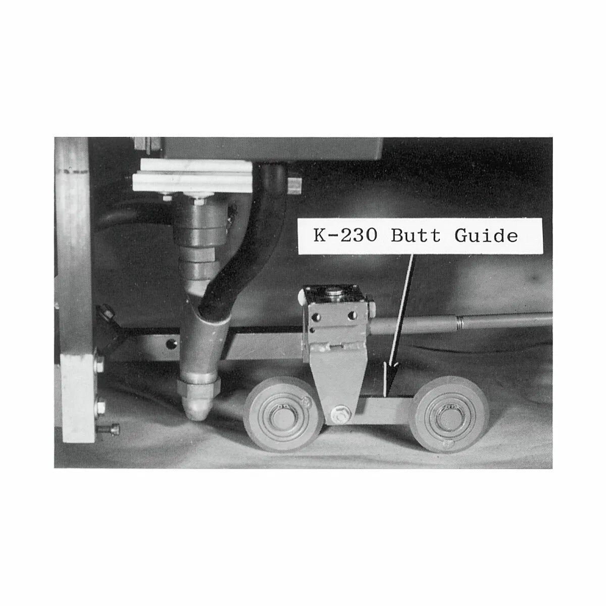 Black and white photo of the Lincoln Butt Seam Guide Kit K230, designed for wire alignment in V-groove welding. The metal part with two axles and wheels is beside a thick pipe with fittings and a downward-pointing nozzle.