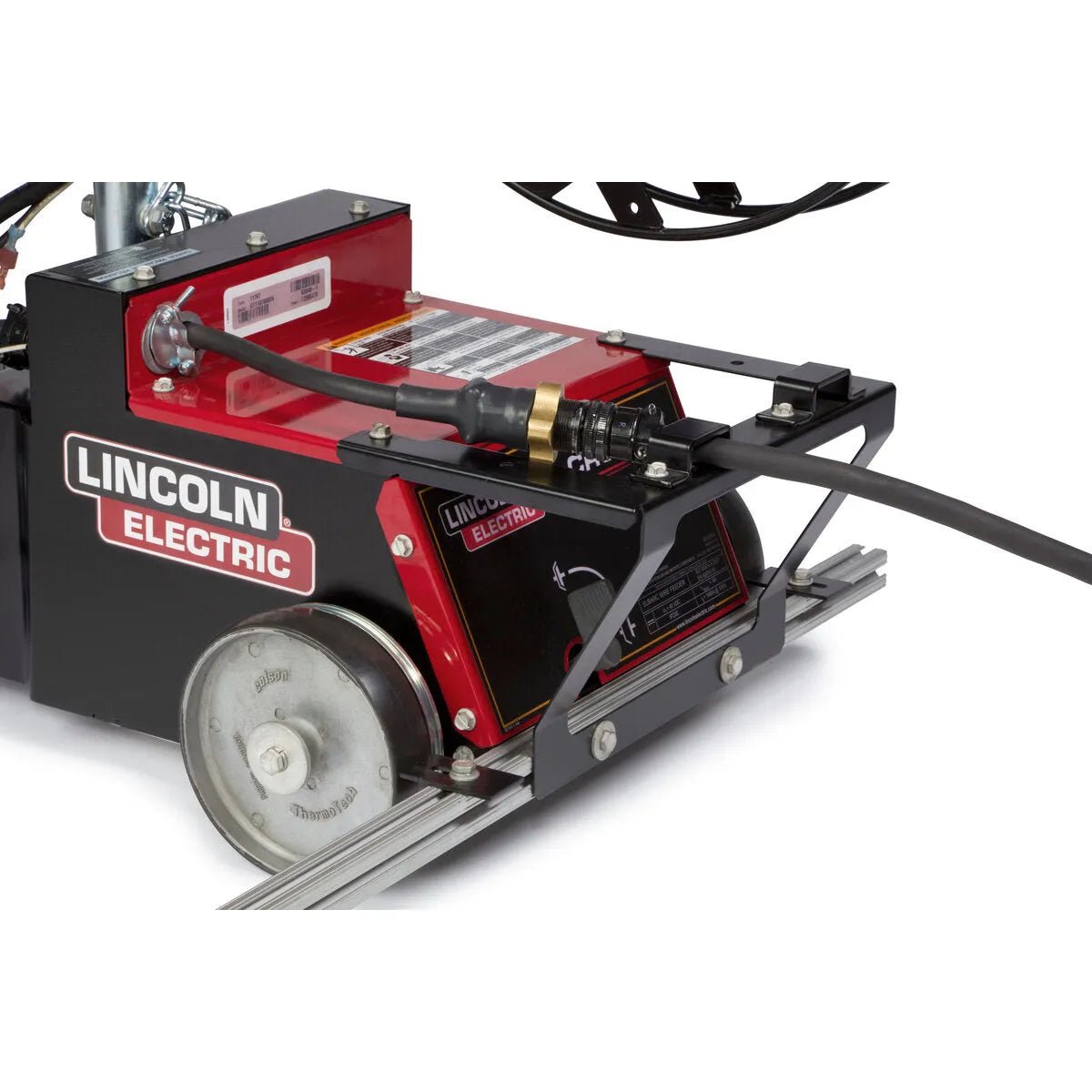 Close-up of a Lincoln Cruiser Submerged Arc Welding Tractor K3048-2 with metallic wheels, black metal frame, thick black cable, barcode sticker, and labels visible as it sits on parallel metal rails.