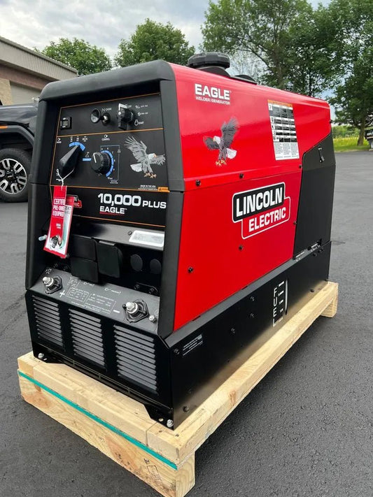 A Lincoln Eagle 10,000 PLUS Factory Demo Engine Driven Welder Kohler (U2343-3) in red and black sits on a pallet outdoors, with its control panel of switches and dials visible. Its set on asphalt, framed by grass, trees, and a parked vehicle.