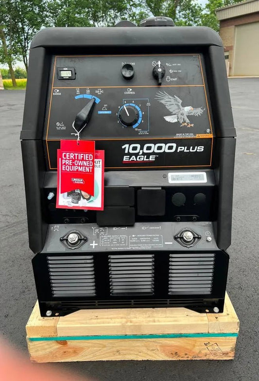 A black Lincoln Eagle 10,000 PLUS Factory Demo Engine Driven Welder Kohler (U2343-3) sits on a wooden pallet outdoors, featuring control knobs, meters, an eagle graphic, and a red certified pre-owned tag. Trees and a building are visible behind.
