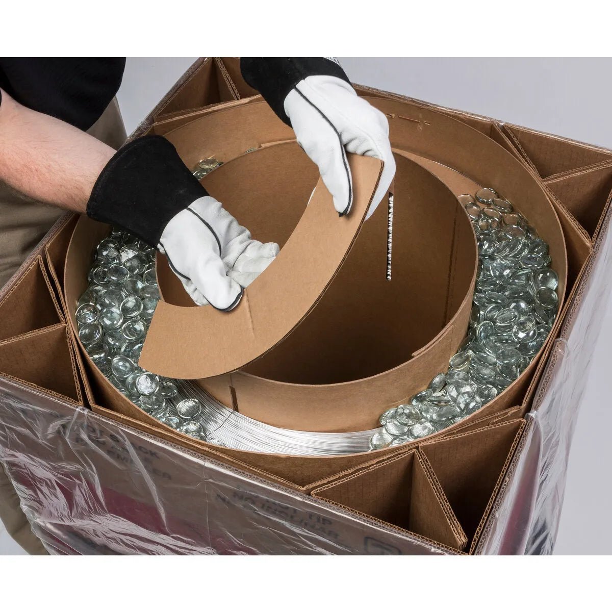 Wearing white gloves with black cuffs, a person lifts a curved Lincoln ED036341 SuperGlaze HD 5183 MIG GMAW Aluminum Welding Wire insert (0.035 in, 300 lb Gem-Pak Box) from a large divided box filled with glass jars and packing material, viewed from above.