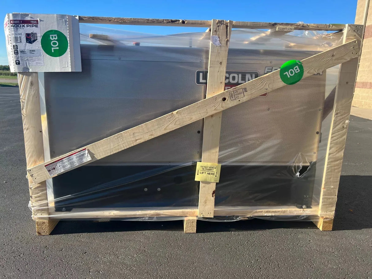 A Lincoln Frontier 400X Pipe Engine Driven Welder Perkins (K3485-1) is wrapped in plastic and crated on asphalt. Shipping label, documents, and green BOL/108 stickers are attached. Blue sky and tan wall seen in background.