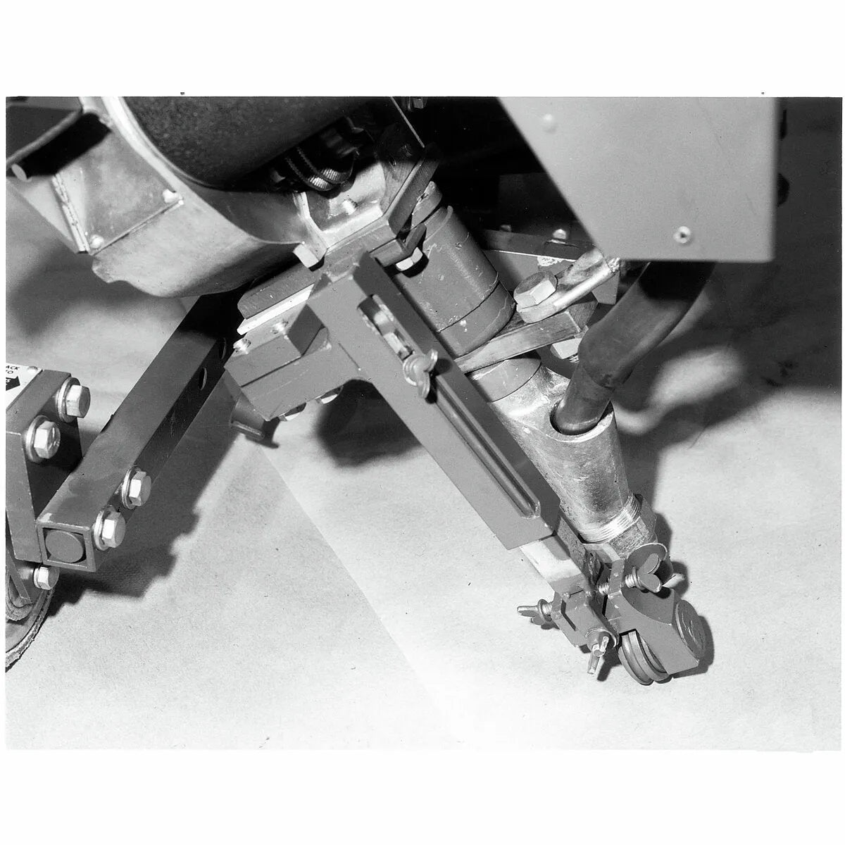 Black and white close-up of a mechanical device, possibly the Lincoln Horizontal Fillet and Lap Adapter Kit K232. The articulated claw-like end effector with wires hovers above a flat surface, emphasizing joints, bolts, and tubing.