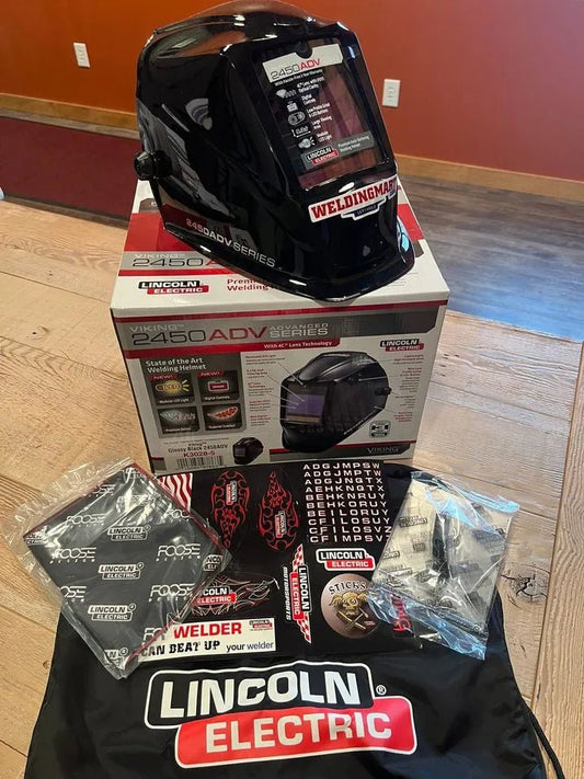 A Lincoln VIKING 2450 ADV Series Black Welding Helmet - K3028-5 with an auto-darkening lens sits on its box on a wooden table, surrounded by stickers, black and red welding gloves, a Lincoln Electric bag, and a sealed manual.