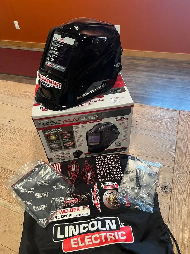 A Lincoln VIKING 2450 ADV Series Black Welding Helmet - K3028-5, with auto-darkening lens, rests on its box amid protective bags, Lincoln Electric decals, a user manual, and replacement lens covers on a wooden table against a brown wall.