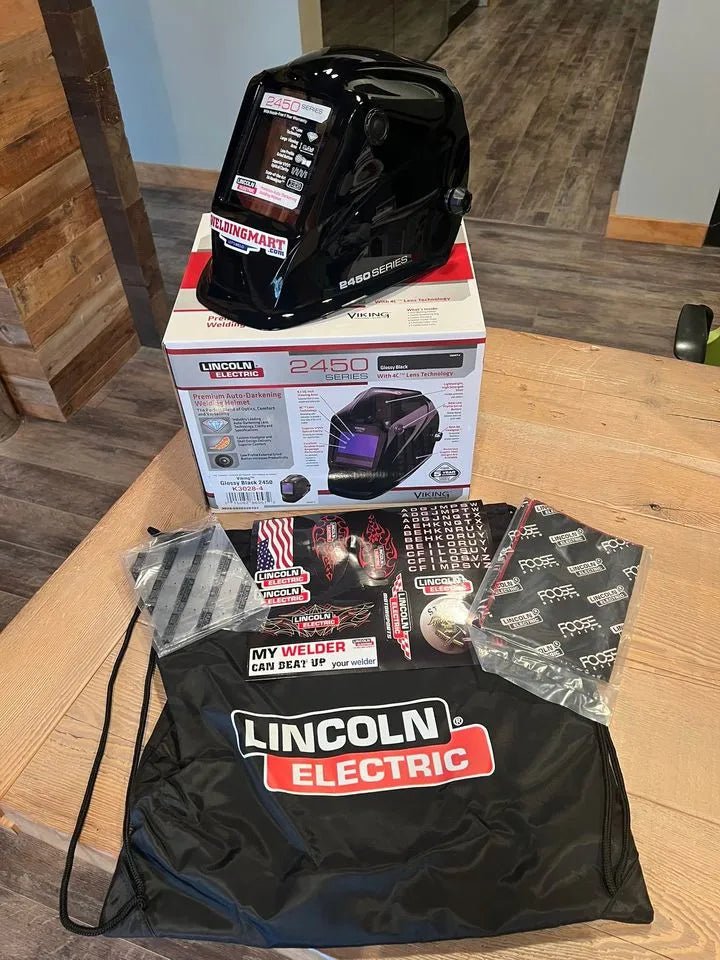 A Lincoln VIKING 2450 Black Welding Helmet - K3028-4 with auto-darkening lens rests on its box on a wooden table, alongside a drawstring bag, two instruction booklets, a sticker sheet, and extra replacement lens covers—all neatly arranged indoors.