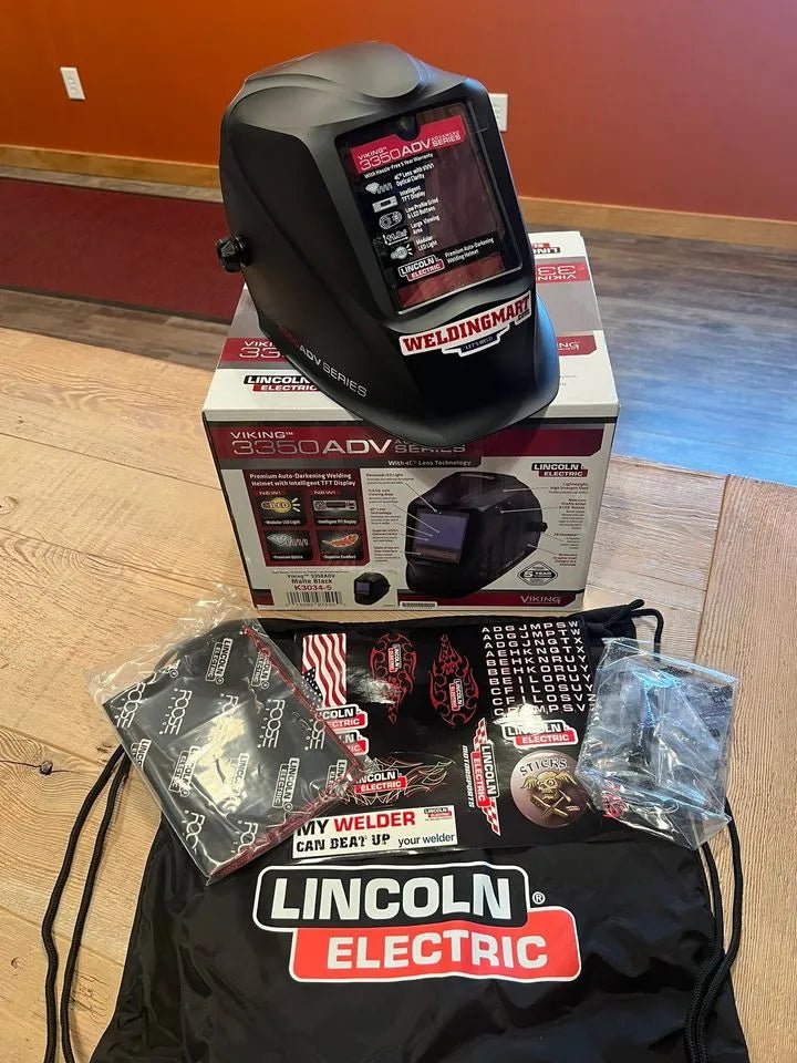 A Lincoln VIKING 3350 ADV Black Welding Helmet - K3034-5 with an auto-darkening lens sits on its box. In front are a drawstring bag, gloves, face mask, stickers, and a sealed clear item on a wood table by a reddish wall.