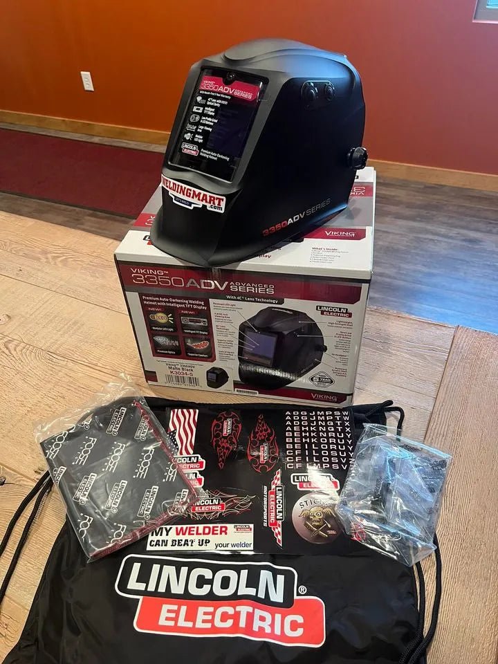 A Lincoln VIKING 3350 ADV Black Welding Helmet (K3034-5) with an auto-darkening lens is displayed on its box, alongside two clear lens covers, a drawstring bag, a sticker, and a patterned bandana on a wooden floor.