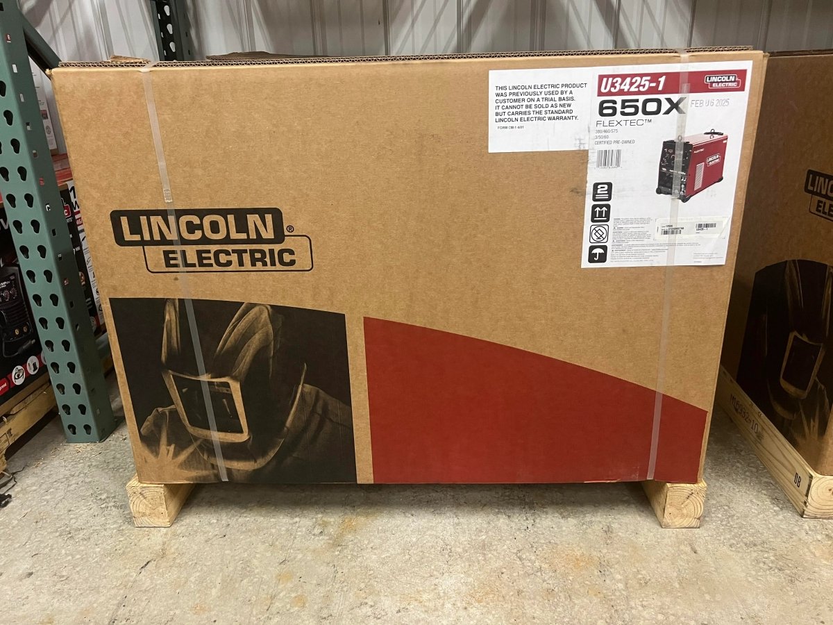 A large box on a warehouse floor contains a Lincoln Flextec 650X Multi-Process Factory Demo Welder with CrossLinc Technology (U3425-1). The box shows the Lincoln Electric logo, a welding mask image, and an upper right product label.