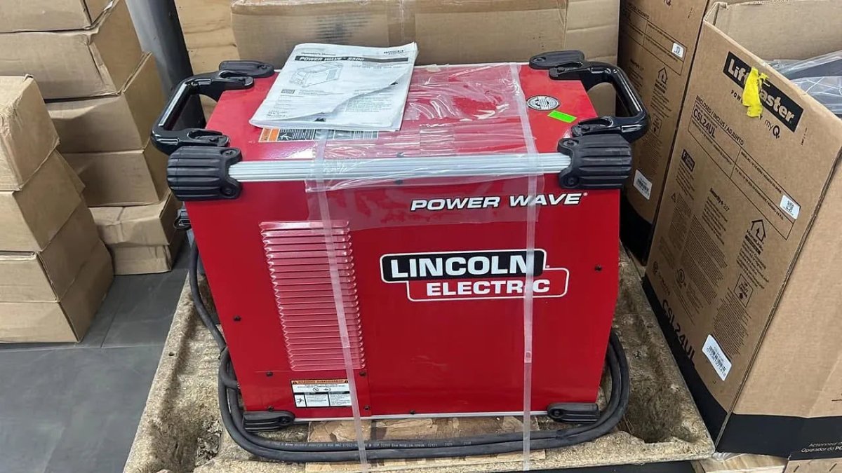 A Lincoln Power Wave S500 Advanced Process Factory Demo Welder (U2904-1) in red sits on a pallet with cardboard boxes, an instruction manual on top, and plastic straps securing the industrial equipment.