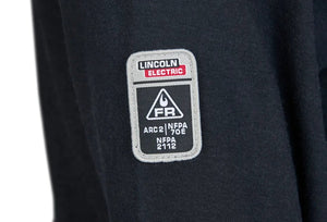 Close-up of the Lincoln K5267-2XL Arc Rated FR Welding T-Shirt (2XL), showing a black fabric sleeve with a rectangular Lincoln Electric patch and safety standards such as ARC 2, NFPA 70E, and NFPA 2112 alongside related symbols.