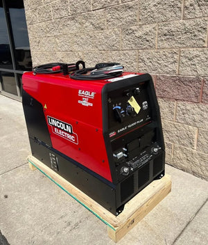 A Lincoln Eagle 10,000 Plus Engine Drive Factory Demo Welder (U2343-4) in red and black sits on a wooden pallet outside, near a tan brick wall and glass window.
