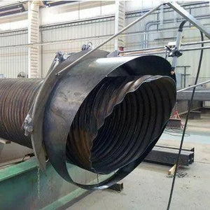 A large industrial pipe with a severely deformed and crumpled end is secured by metal straps in a factory or workshop setting. The damaged pipe appears to have been crushed or twisted.