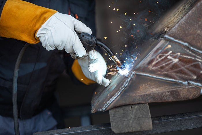 A person wearing protective gloves and a yellow safety sleeve welds two metal pieces together. Bright sparks fly from the welding torch, illuminating the metals surface. The person’s face is not visible, focusing on their hands and the welding action.