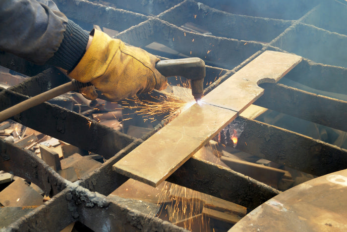 A person wearing protective yellow gloves uses a plasma torch to cut a sheet of metal on a grid table. Bright sparks fly from the cutting line, and some smoke rises around the area, indicating active metalworking in an industrial setting.