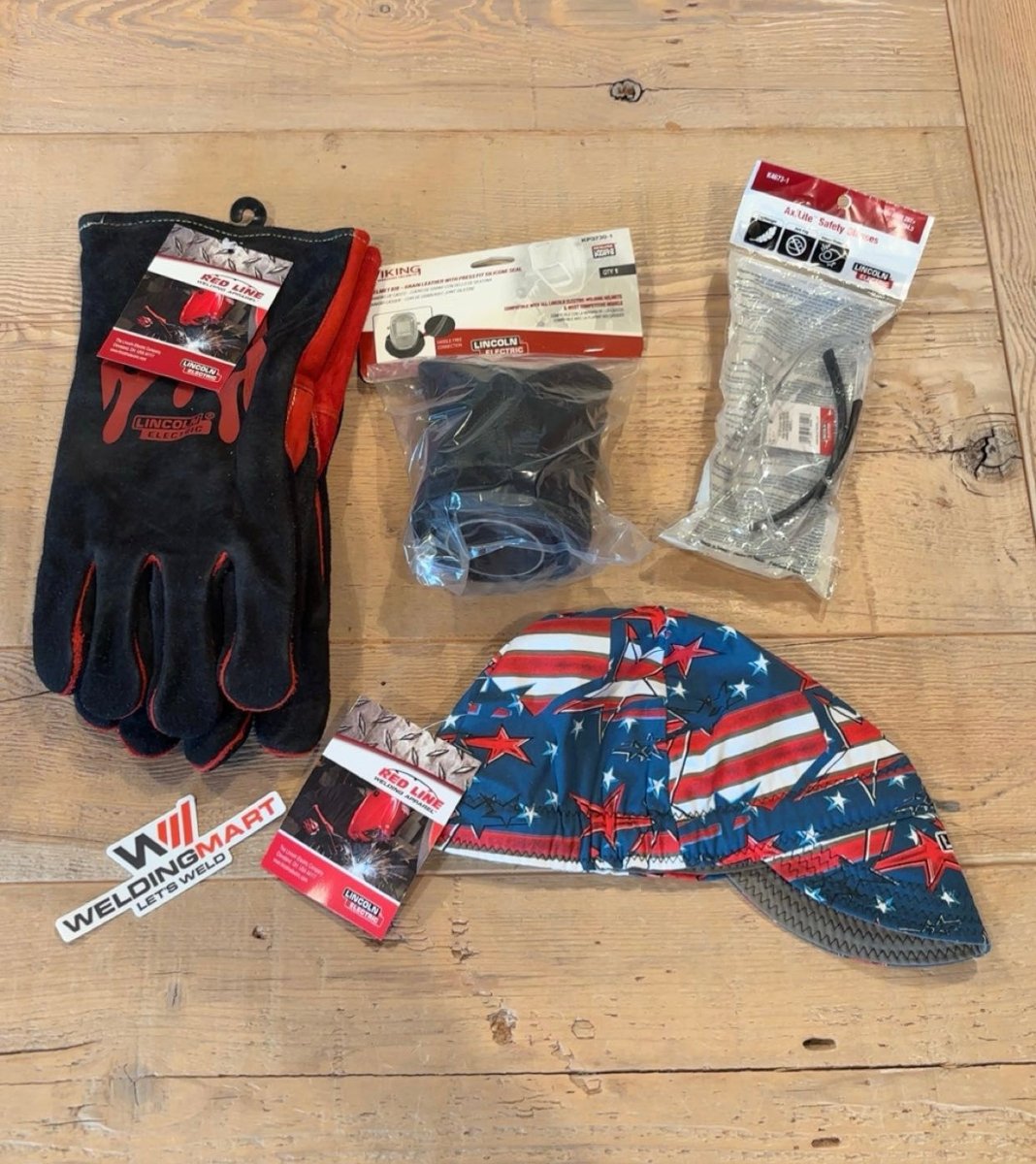 WeldingMart Lincoln Viking Helmet Bonus: welding gloves, goggles, cap, cable ties, sticker, and lenses on wood surface.