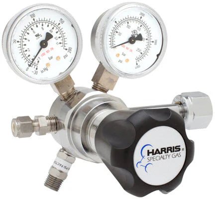 A close-up of the Harris Regulator 721C-050-350-F (721C050350F) shows two pressure gauges, a black knob labeled Harris Specialty Gas, and metallic fittings. This high-purity regulator has a polished finish for connecting gas lines.