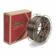 A spool of Lincoln .052 METALSHIELD MC-706 33# SSP ED031584 welding wire with a metallic finish sits before a red and brown box featuring the Lincoln Electric logo and slogan, THE WELDING EXPERTS. The coiled wire and metal supports are visible.