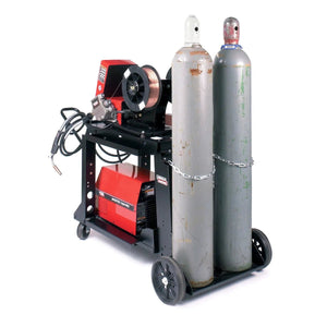 A black cart with the Lincoln Dual Cylinder Mounting Kit K1702-1 secures two large gas cylinders by chain and holds a red welder, wire feeder, hoses, and cables—ideal for efficient MIG welding setups.