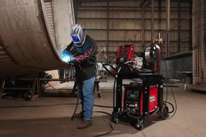 Wearing a welding helmet and gloves, a worker welds a large metal structure indoors as bright sparks fly near a red Lincoln Inverter and Wire Feeder Cart K1764-1 with attached cables in a spacious, well-lit industrial area.