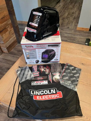 A Lincoln VIKING 2450 Black Welding Helmet - K3028-4 with auto-darkening lens rests on its box on a wooden table, alongside a drawstring bag, two instruction booklets, a sticker sheet, and extra replacement lens covers—all neatly arranged indoors.