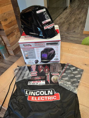 A Lincoln VIKING 2450 Black Welding Helmet - K3028-4 with an auto-darkening lens is displayed on its box, surrounded by a drawstring bag, safety gloves, stickers, a neck gaiter, and info cards. Wood and tile floors are visible indoors.