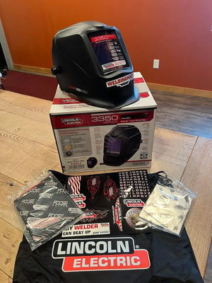 A Lincoln VIKING 3350 Black Welding Helmet - K3034-4 with auto-darkening lens rests on its box atop a wooden table, surrounded by instruction booklets, decals, and a Lincoln Electric protective bag. The helmet display shows model 3350.