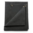 The Lincoln Welding Blanket - 6 ft x 6 ft - 1100° C Rated (K3254-1) is a black fiberglass blanket with reinforced edges and two metal grommets in the corner. One corner is lifted to display the underside and stitching; it’s neatly stacked on a white background.